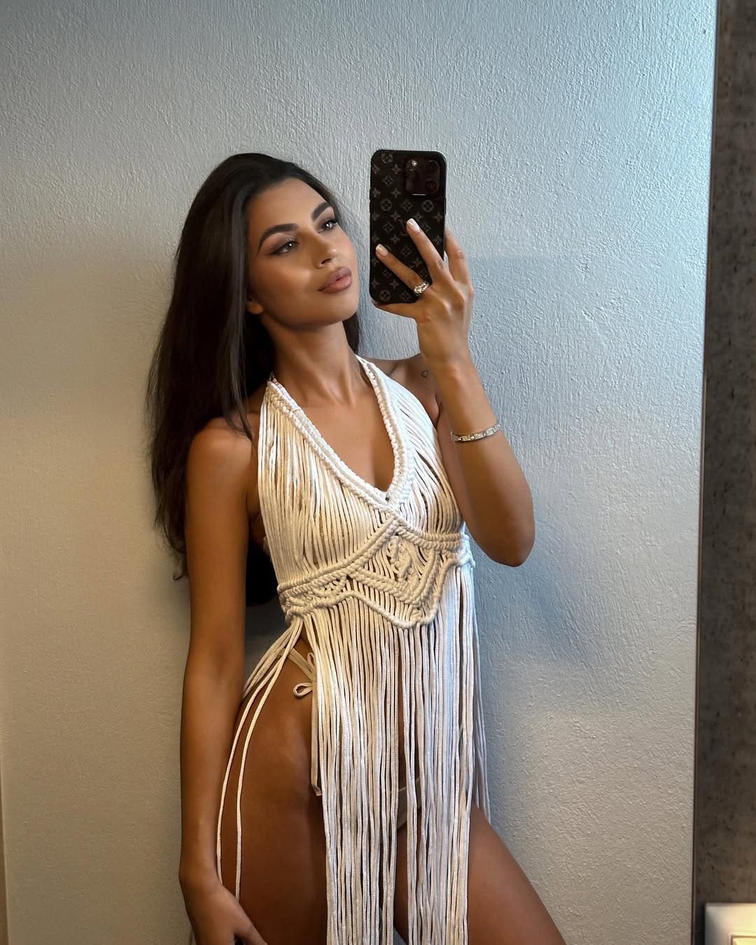 A mirror selfie shows a woman in a white macrame fringe dress with a deep V-neck and open sides, standing against a light wall.