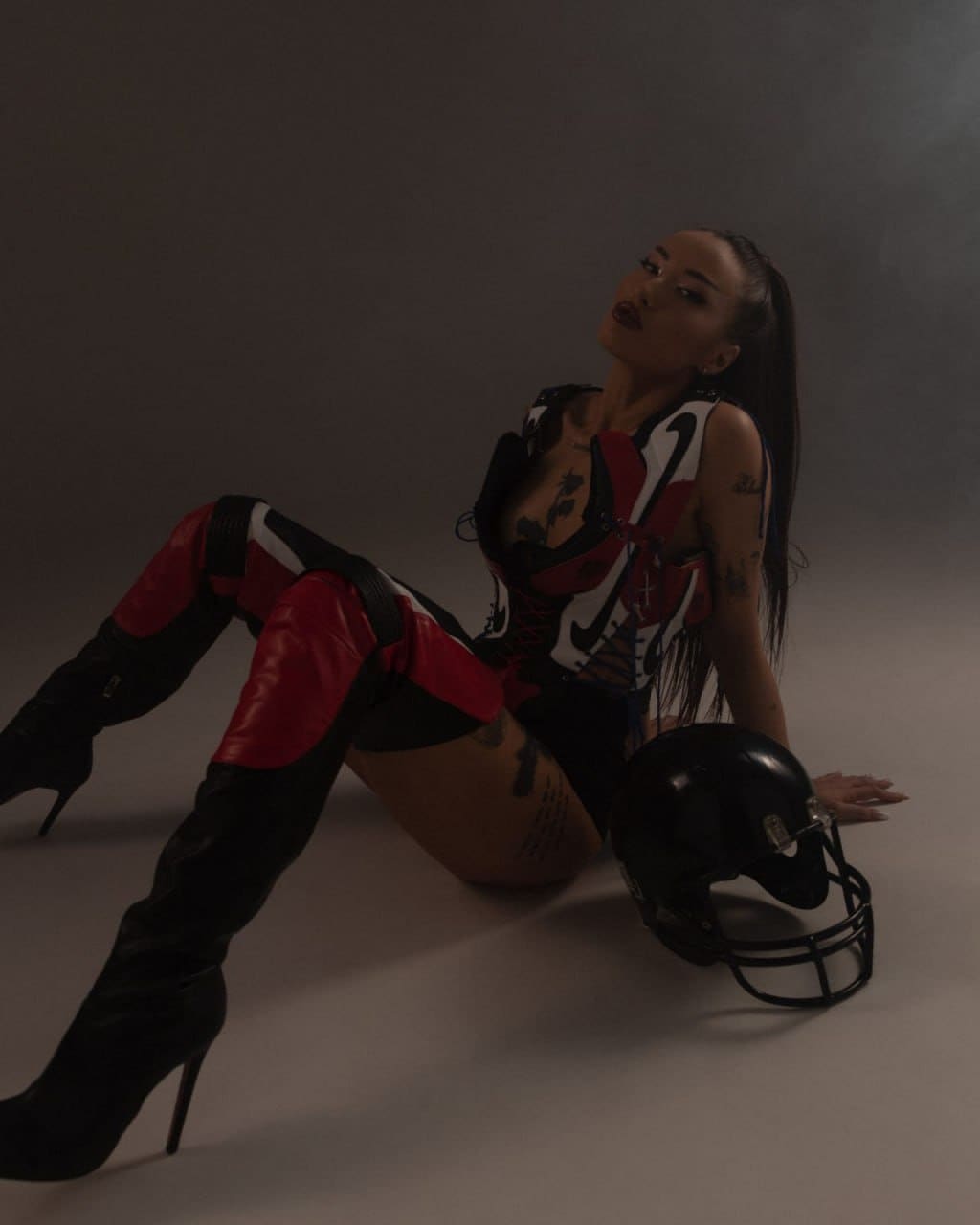 A woman sits on a grey studio floor in a patterned corset, shorts, and thigh-high boots, with a black football helmet beside her.