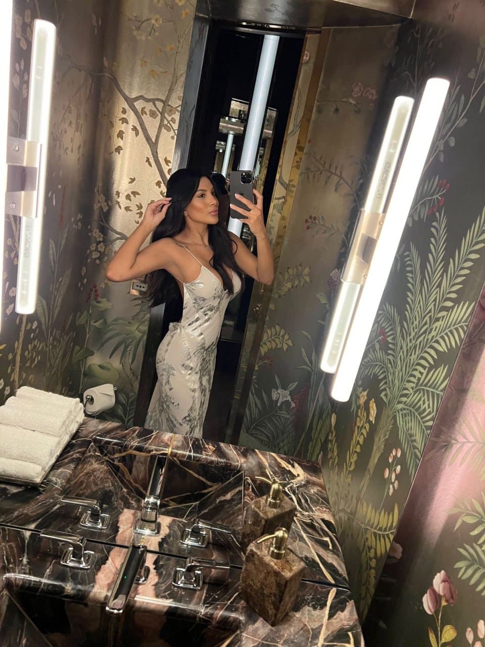 A woman takes a mirror selfie in a bathroom with floral wallpaper, wearing a patterned slip dress, with a marble sink and towels in the foreground.