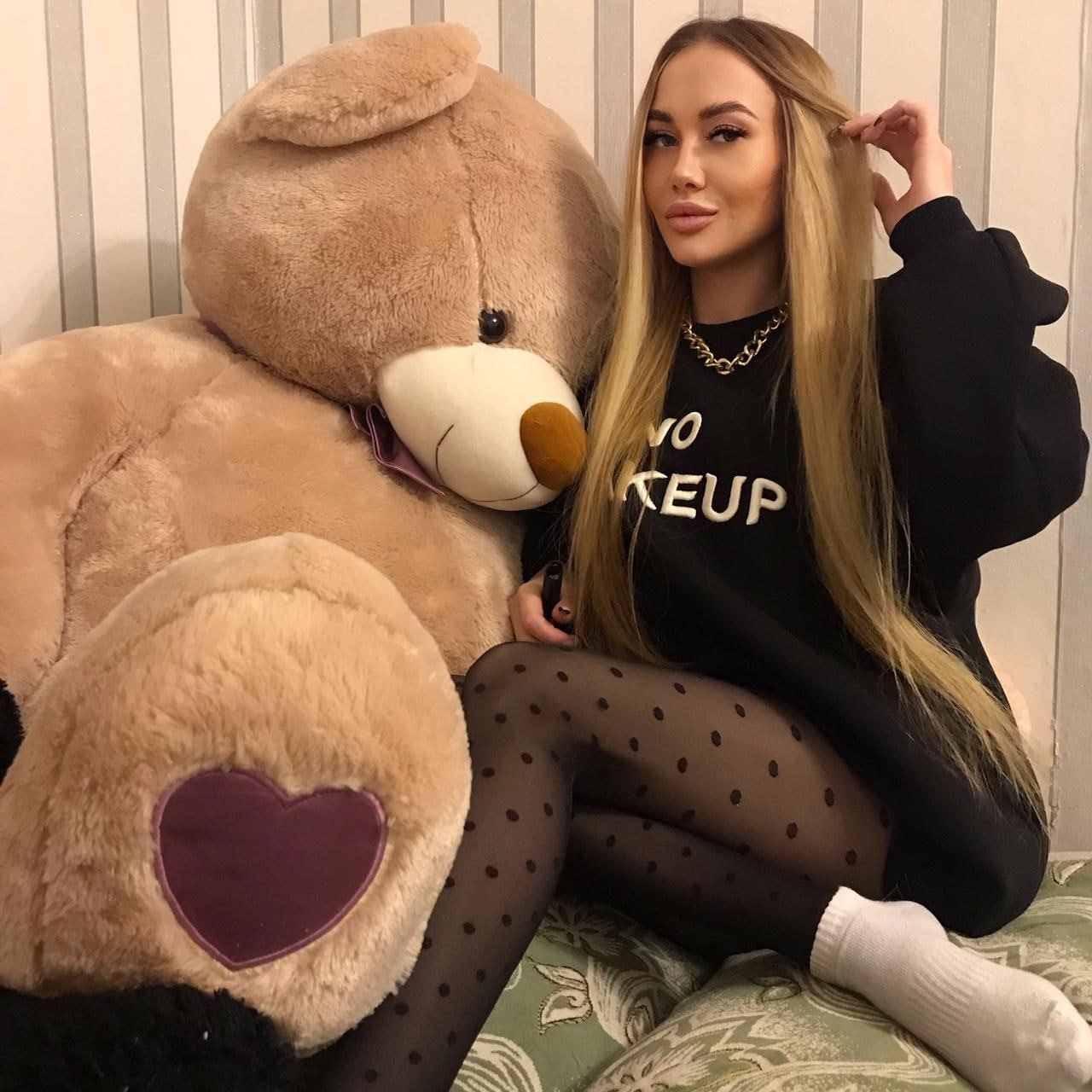 Avgustina sits on a patterned couch beside a tan teddy bear, wearing a black long-sleeved top, polka-dot tights, and white socks.