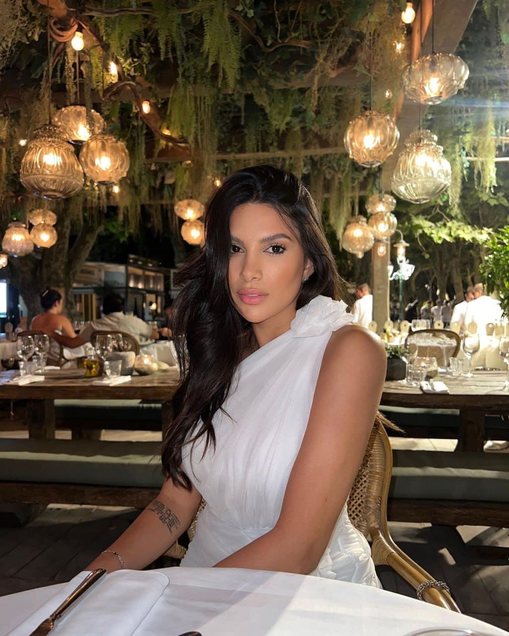 A woman sits at a white-clothed table in a restaurant with hanging lanterns, wearing a sleeveless white one-shoulder dress with a ruffled neckline.
