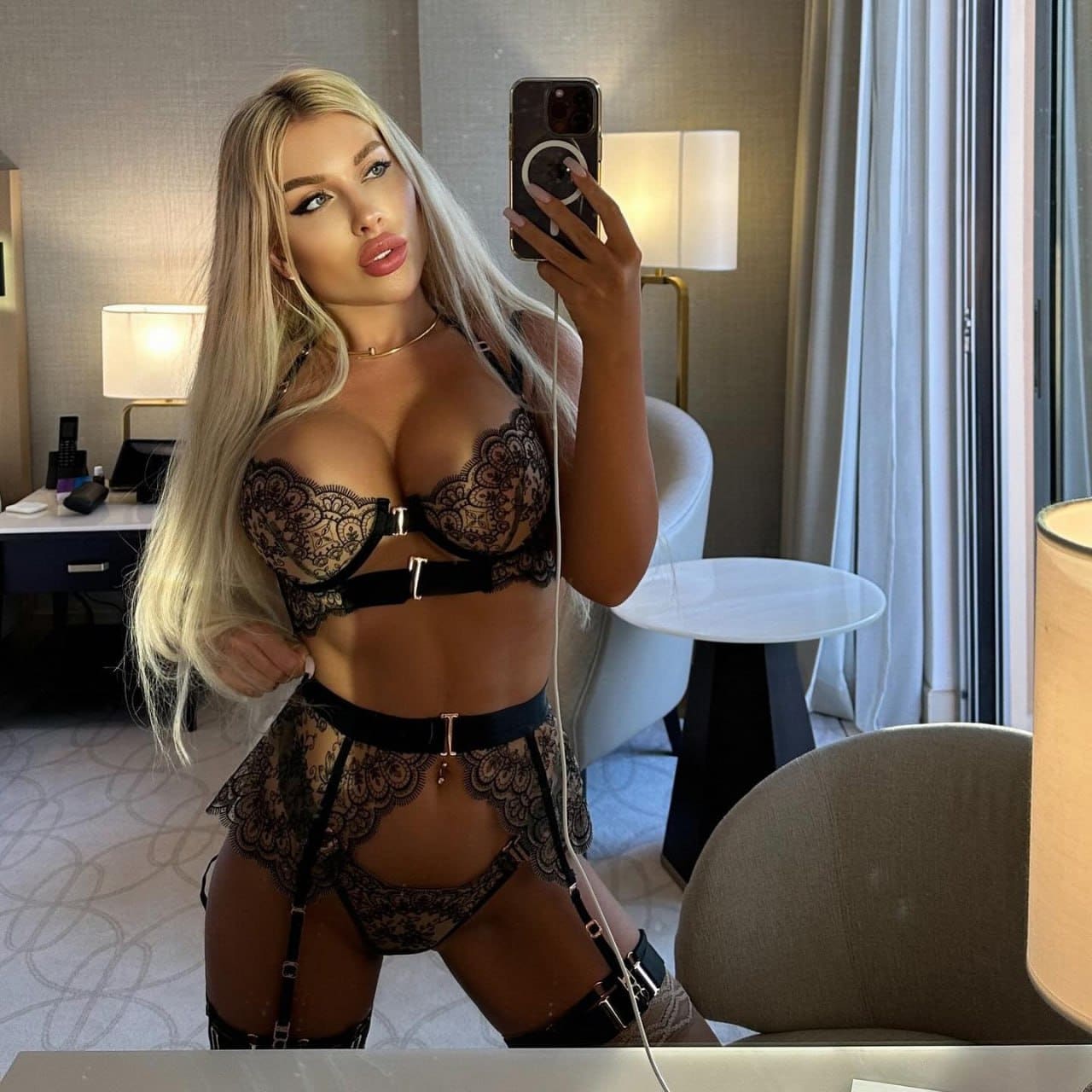 A woman holds a smartphone in a hotel room with a desk and lamps, wearing a black lace lingerie set with garter straps.
