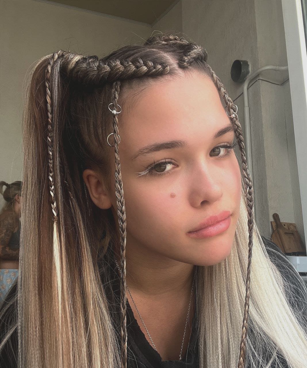 Jenia with braided highlighted hair in a ponytail wears a dark top and necklace, standing indoors by a window against a neutral wall.