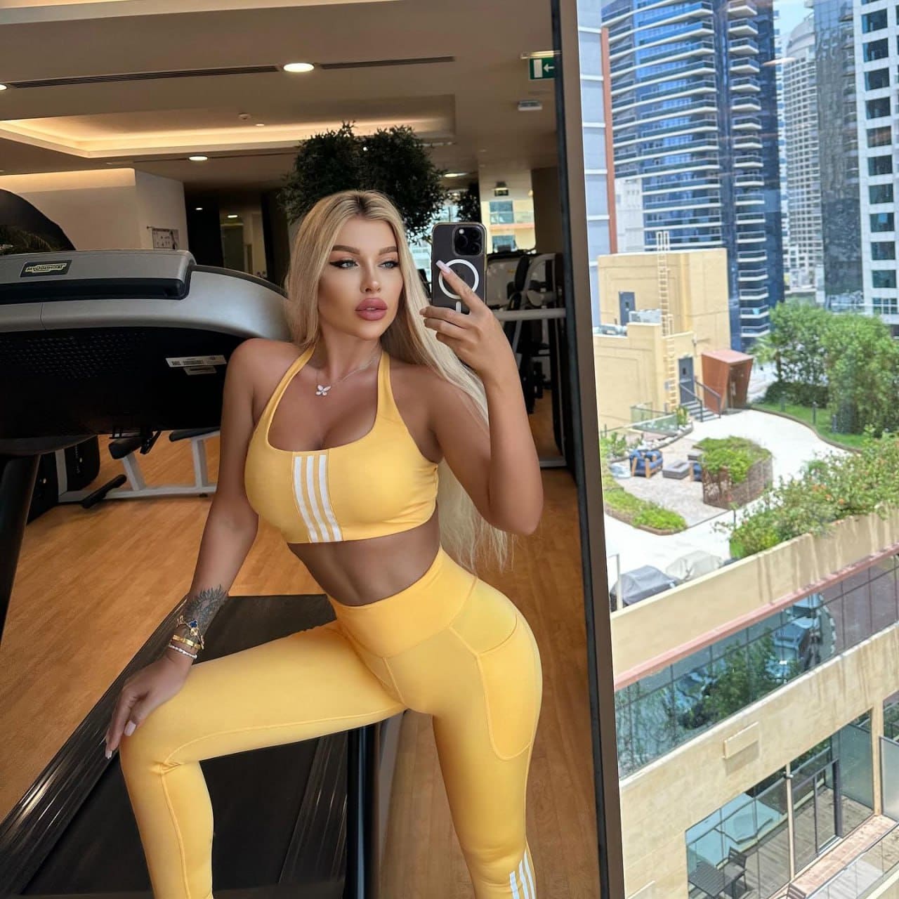 Iskra stands in a gym with a treadmill and city view, wearing a yellow sports bra and leggings while holding a phone.