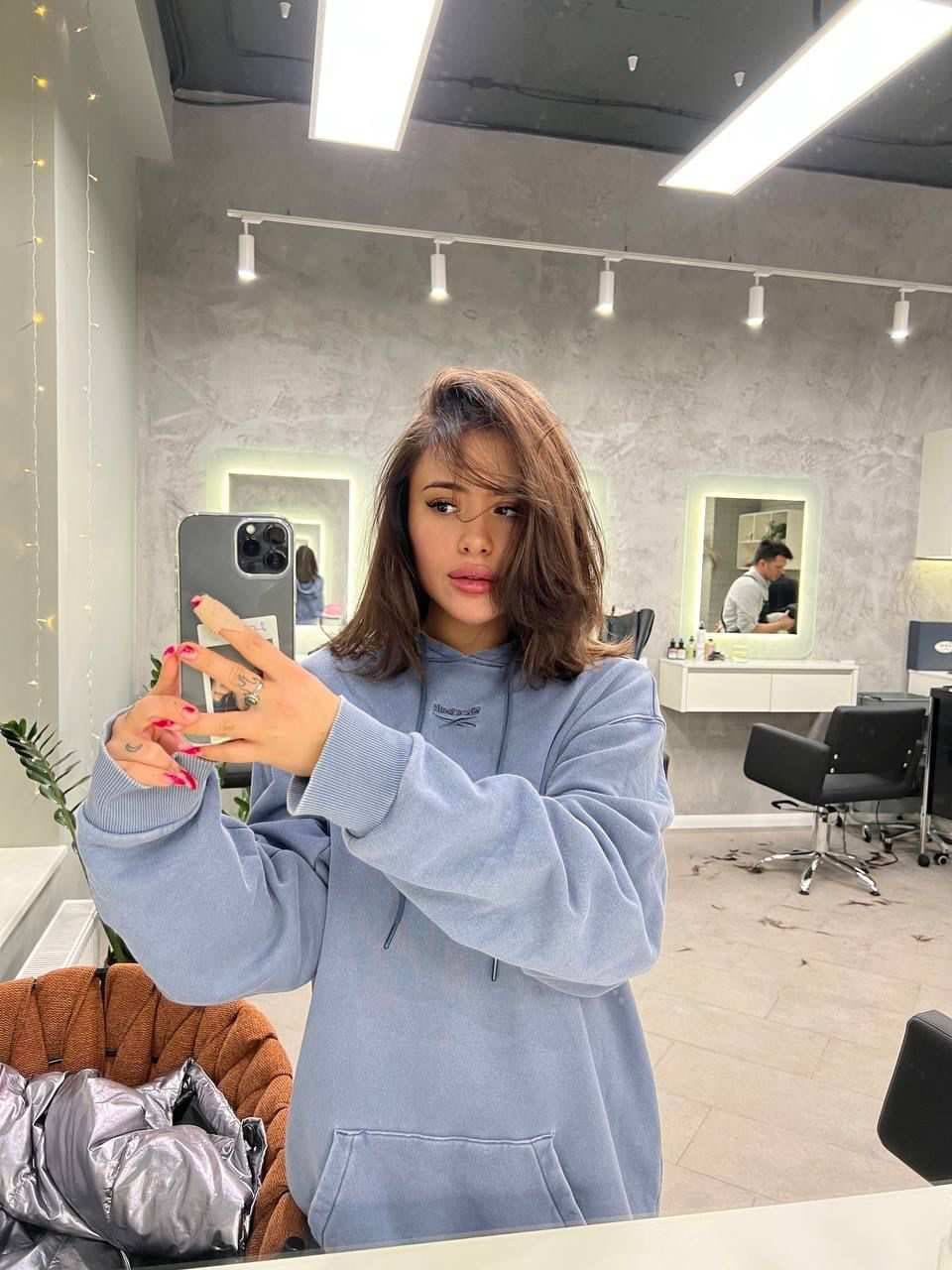A woman in an oversized light blue hoodie takes a mirror selfie in a salon, holding a grey smartphone with red nails.