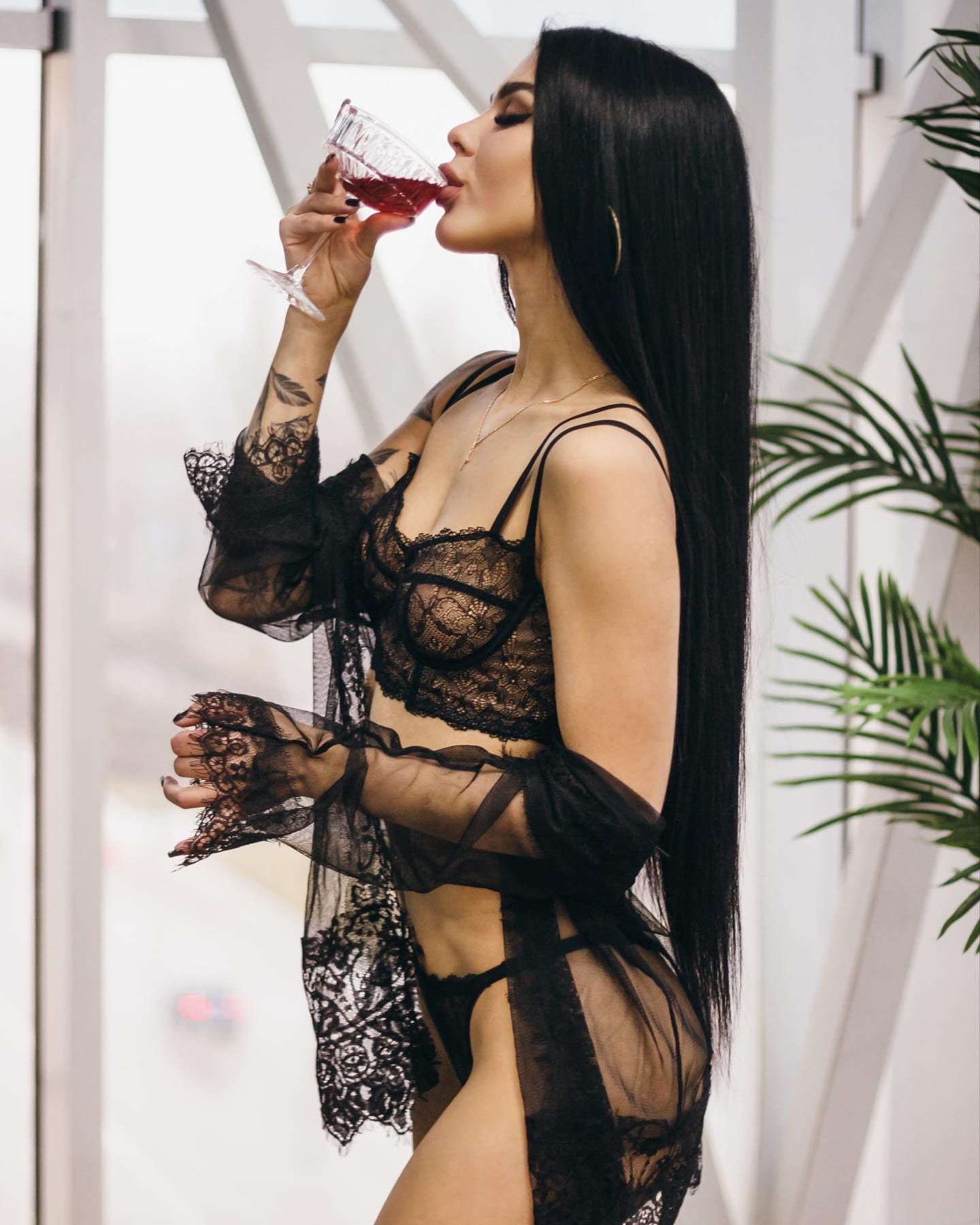 A woman in profile drinking red wine, wearing a black lace bra, garter, and sheer robe, with a forearm tattoo and a potted plant behind.