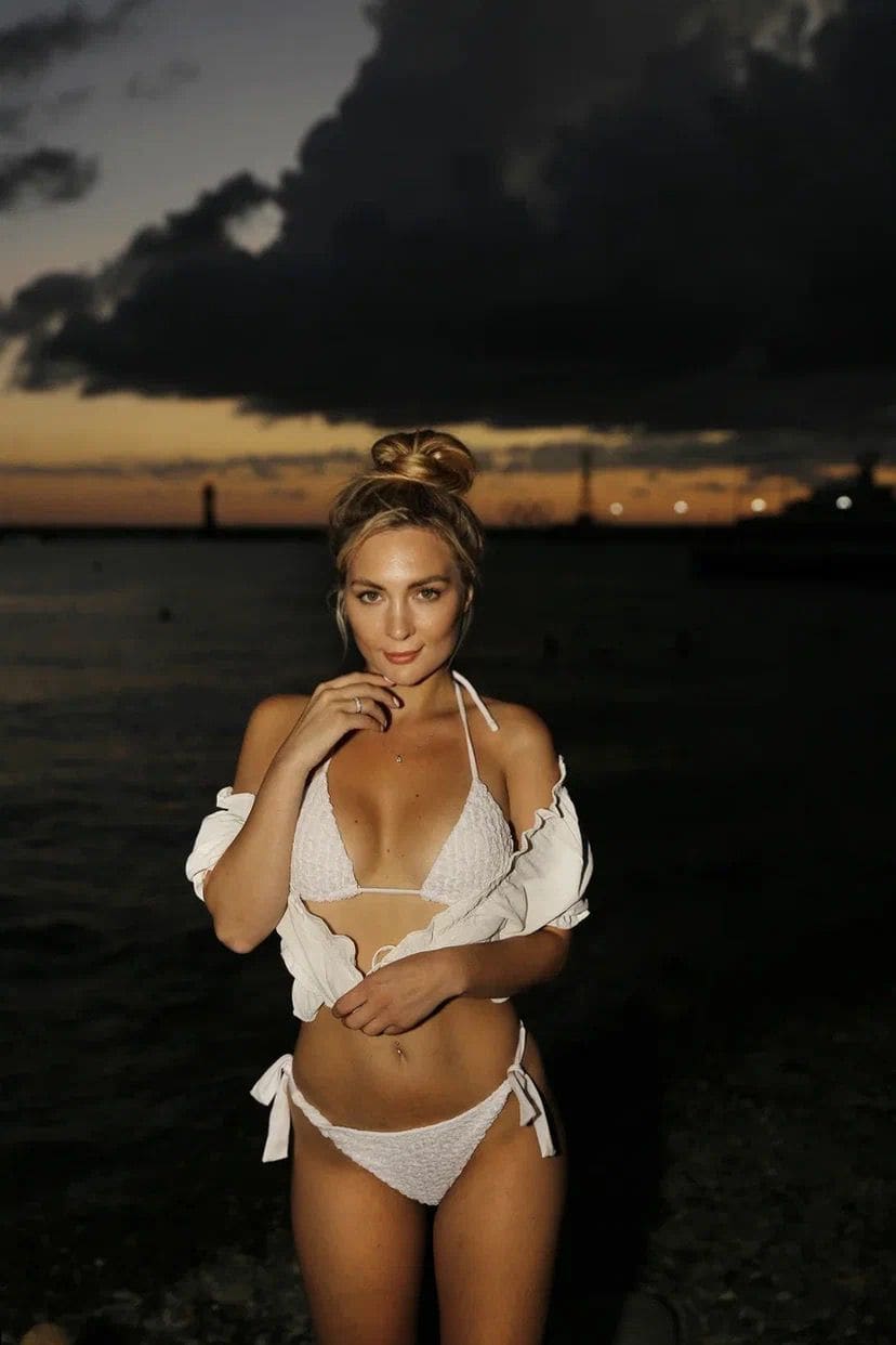 A blonde woman in a white bikini and open shirt poses on a beach at dusk, hand near chin, against dark water and cloudy skies.