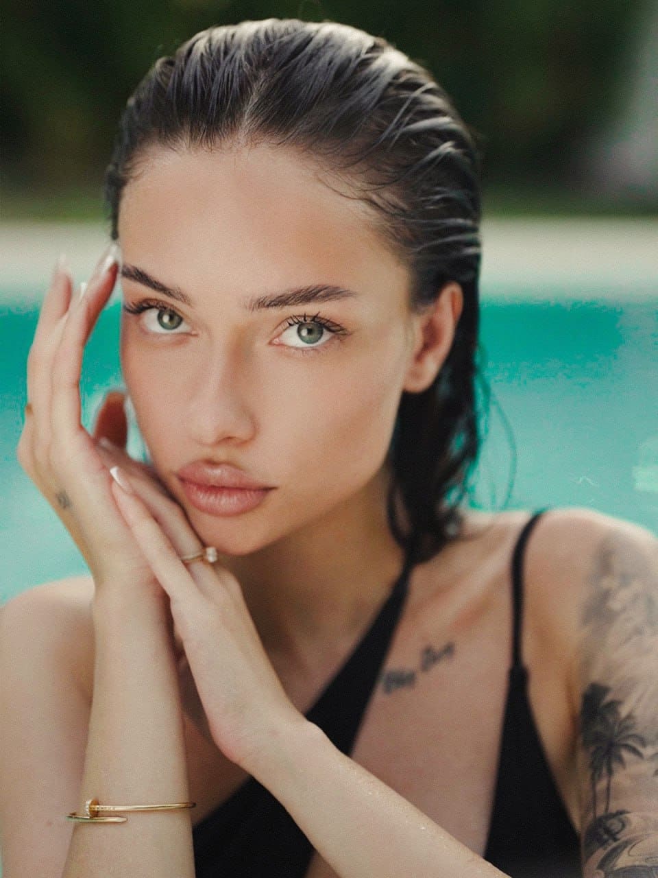 Flora rests her chin on her hands in a black halter top, with a shoulder tattoo visible against a blurred turquoise pool background.