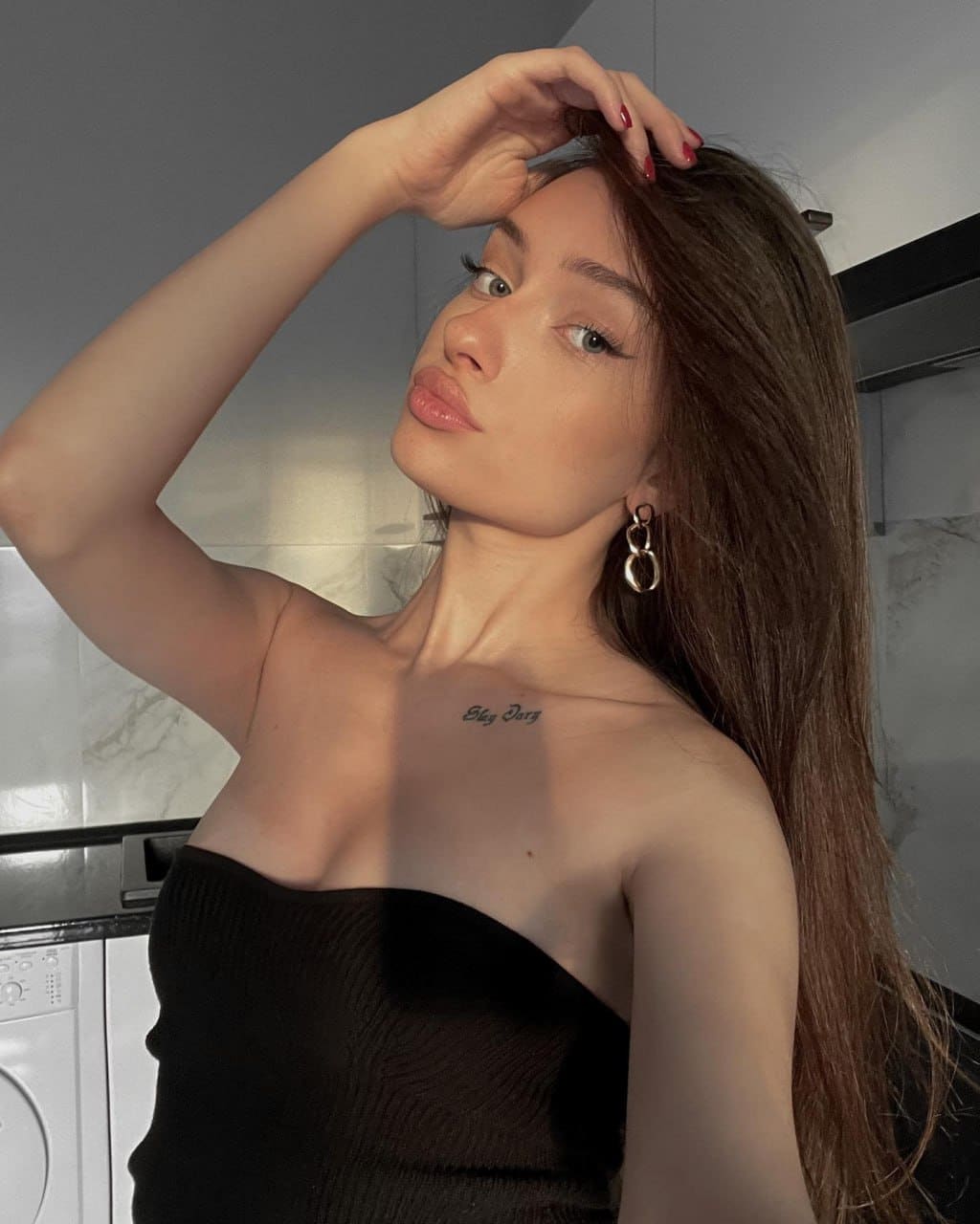 A woman with long brown hair and a chest tattoo poses in a black strapless top, one hand to her head, in a room with marble walls.
