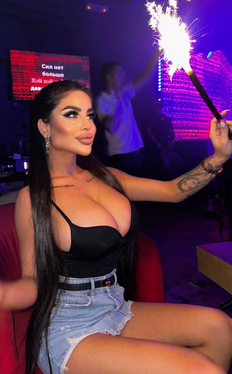 A woman in a black top and denim shorts sits on a red club couch holding a lit sparkler, with a man in the background.