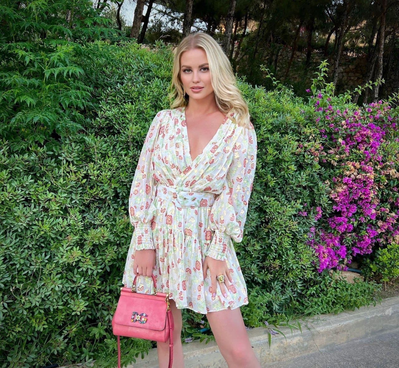 Helen stands before a green hedge and purple flowers, wearing a white floral wrap dress and holding a small pink handbag.