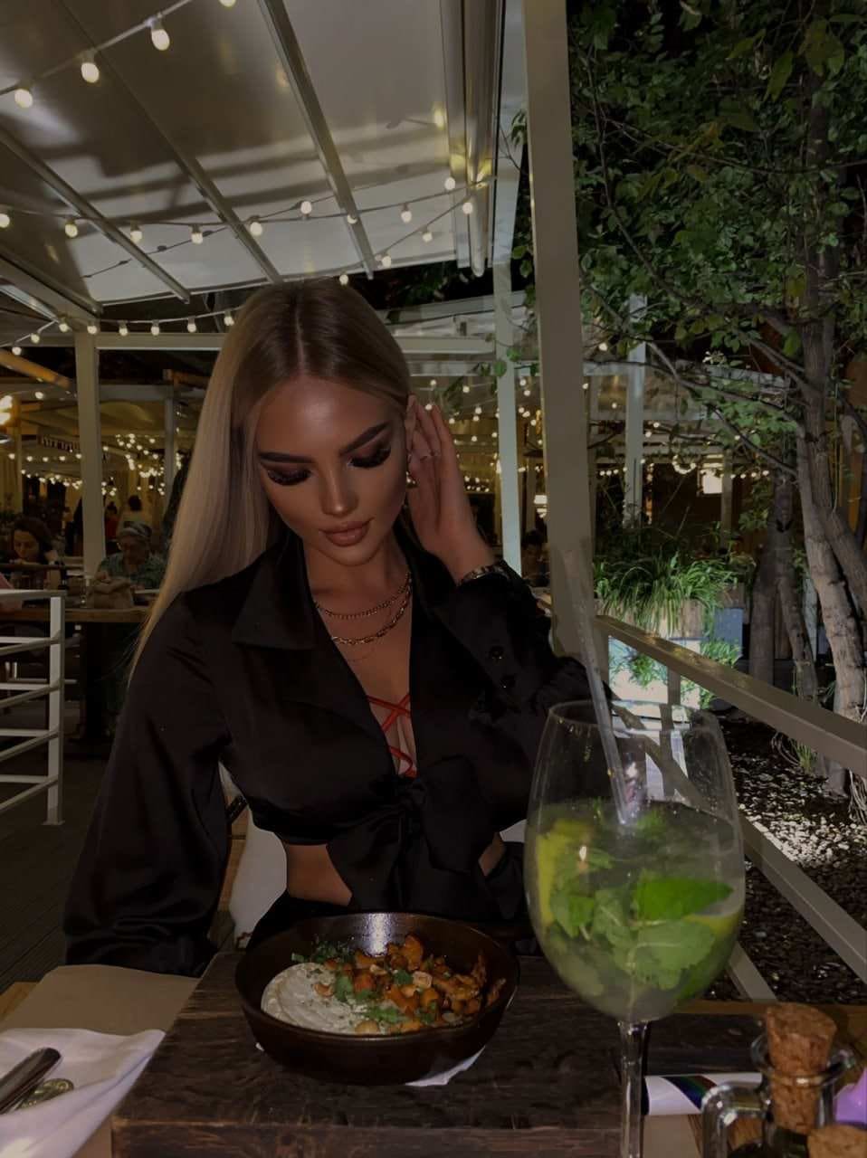 Adeline sits at an outdoor table at night in a black tied shirt over a red top, with food and a drink under string lights.