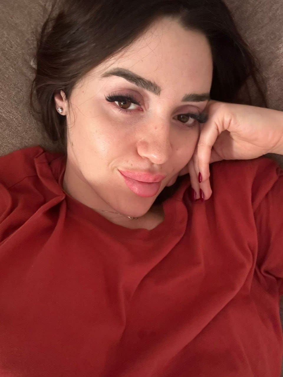 A woman lies on a textured gray surface in a loose red top, resting her head on a hand with red-painted nails.