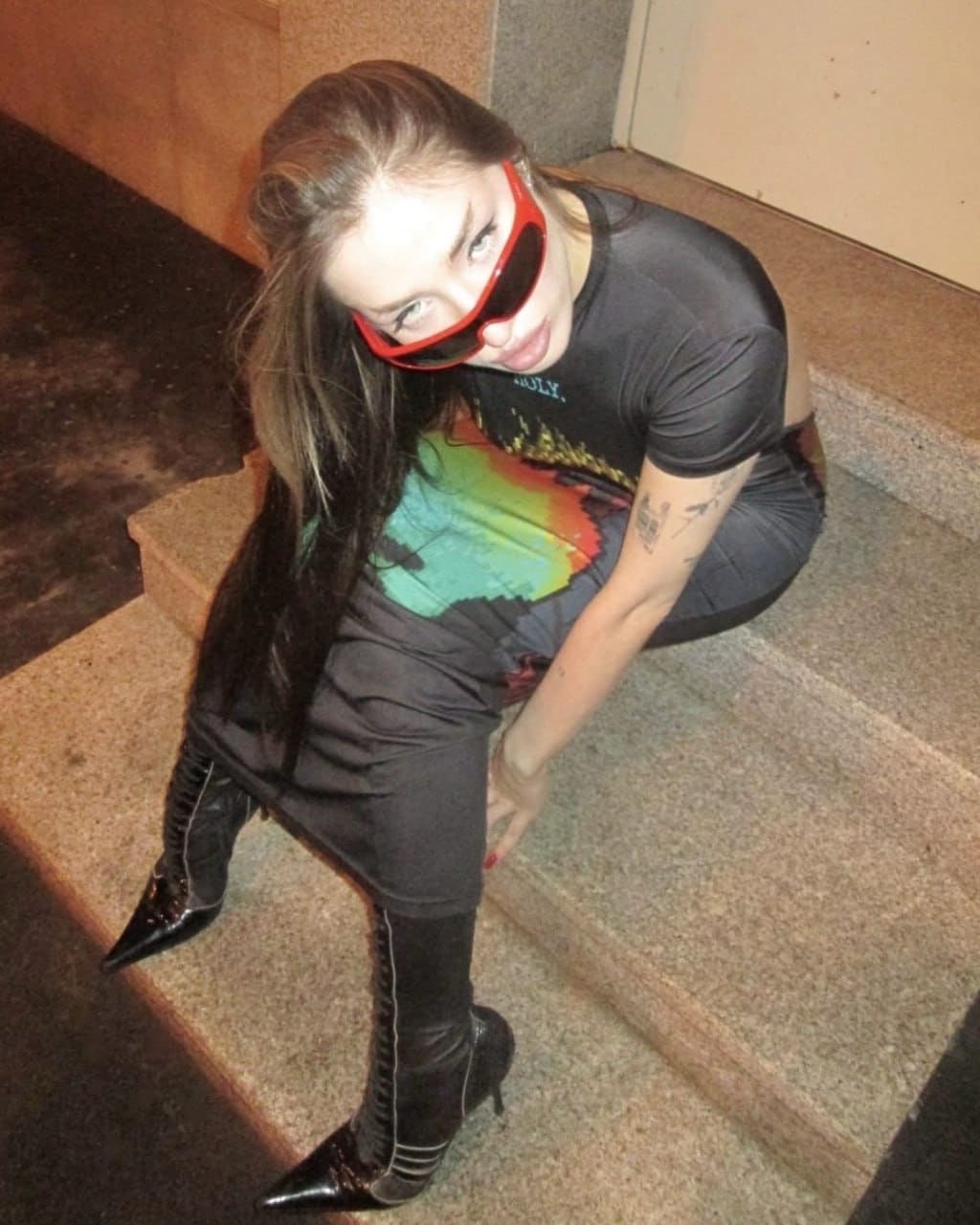 A woman sits on stone steps wearing a graphic t-shirt, black leggings, lace-up boots, and red sunglasses, with a forearm tattoo visible.