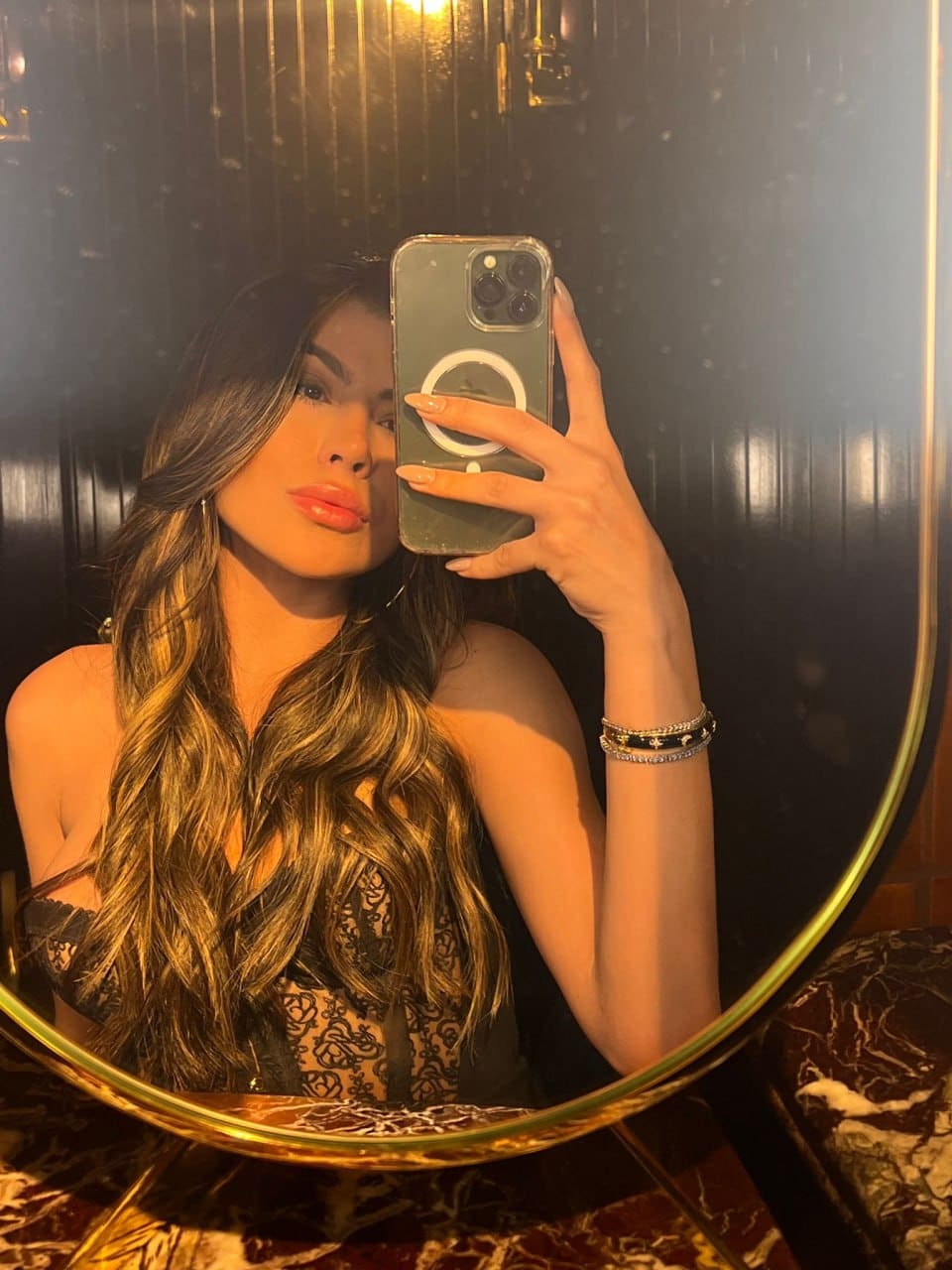 A mirror selfie captures a woman in a black lace top with bare shoulders in a dimly lit room.