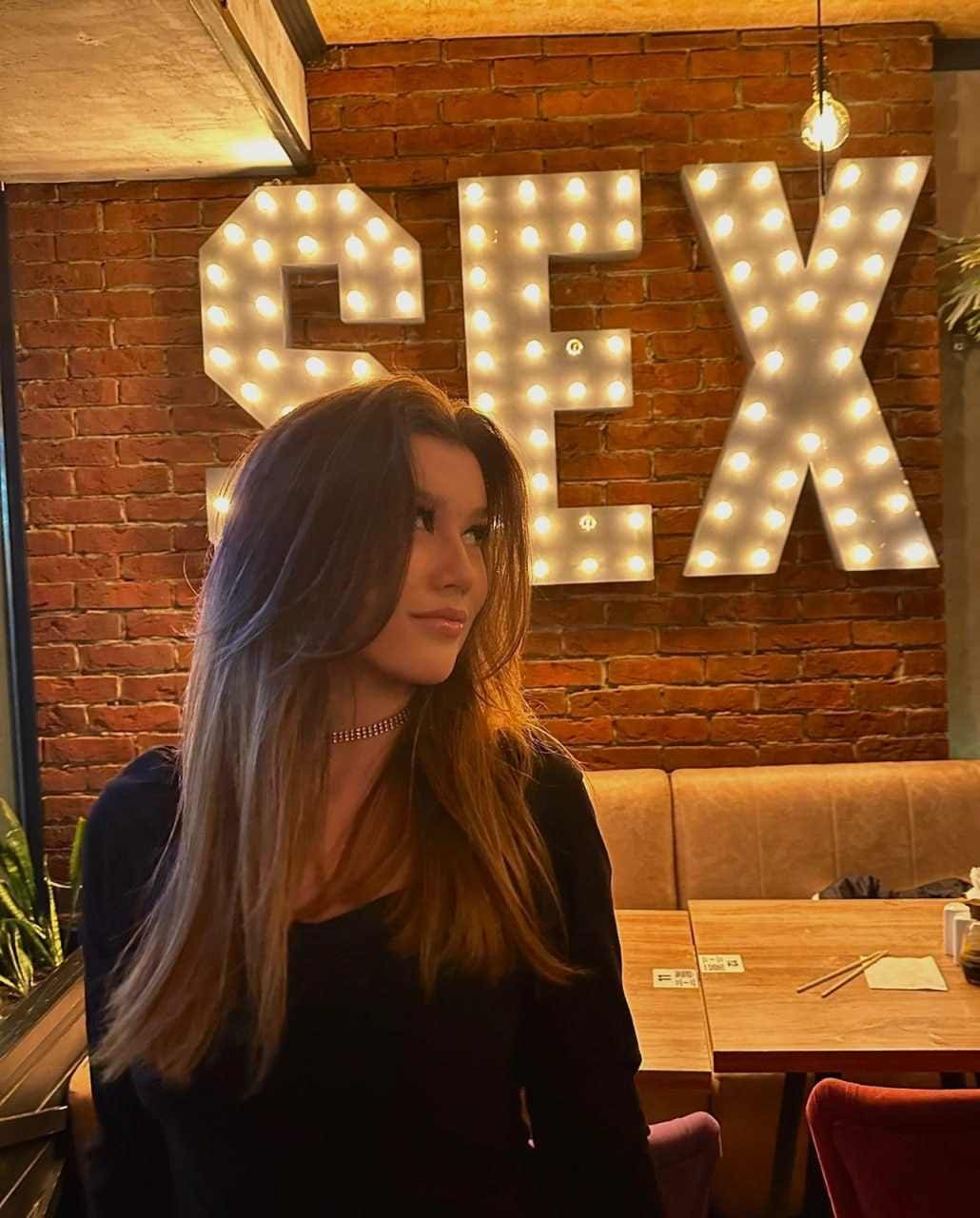 Lena sits in a restaurant wearing a black long-sleeved top and beaded choker, with a brick wall and lit 'SEX' marquee letters behind her.