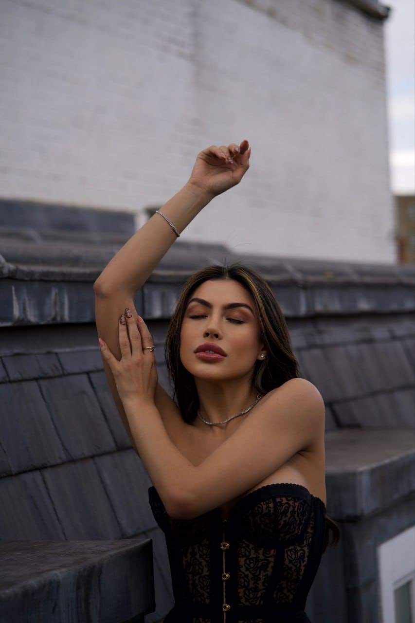A woman stands on a rooftop in a black lace strapless top with one arm raised against a brick wall.
