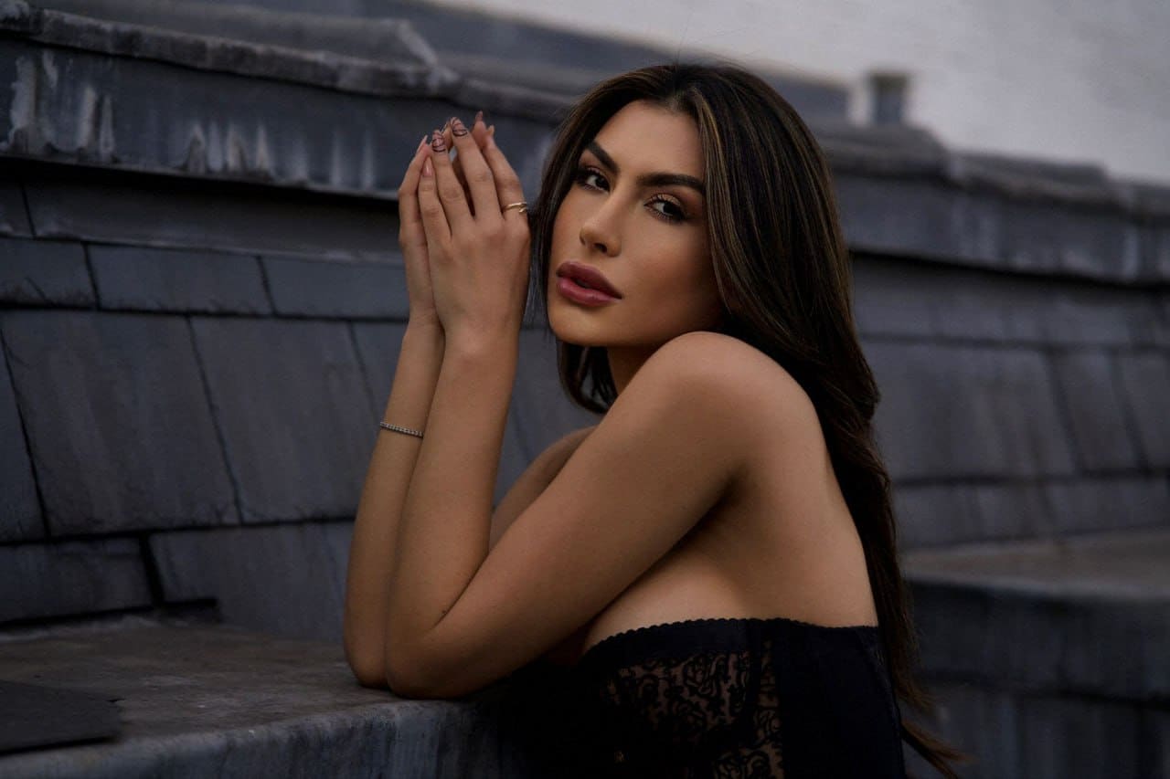 A woman sits on stone steps wearing a black lace off-the-shoulder top with hands clasped near her face.