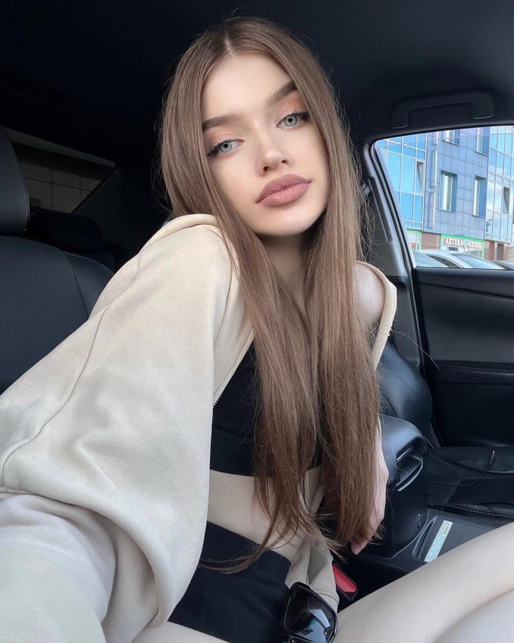 Sitting in a car's back seat, she wears a cream hoodie over a black top, with sunglasses on her lap and a city street outside.