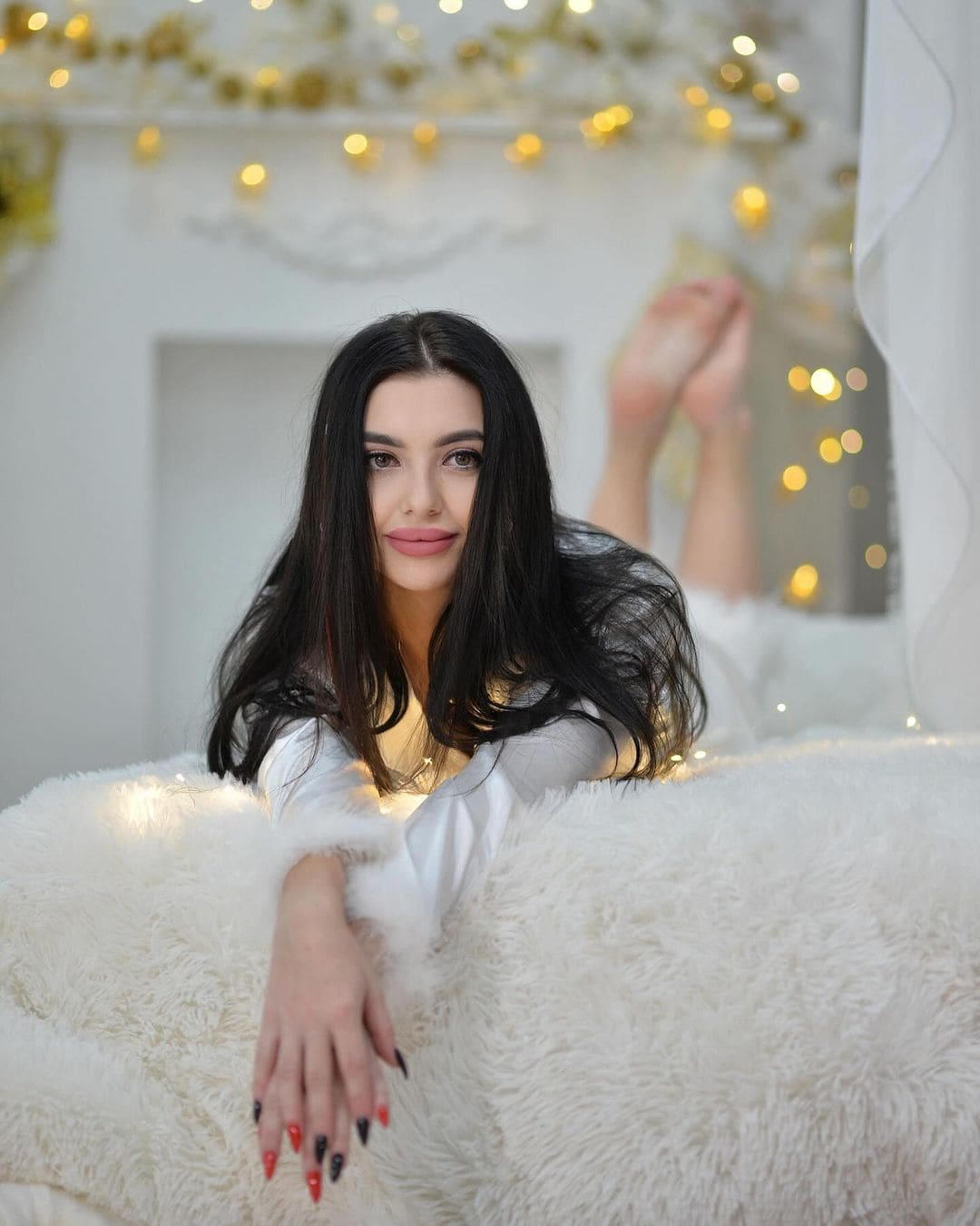A woman in a white long-sleeved top lies on her stomach on a fluffy white surface, feet raised, with string lights behind.