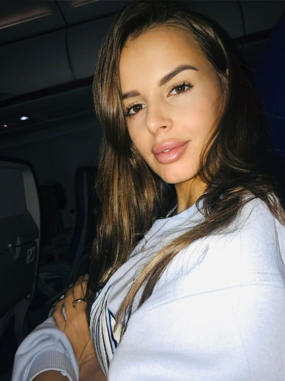 Seated inside a vehicle, she wears a light blue hoodie over a striped top, with one hand resting near her chest.