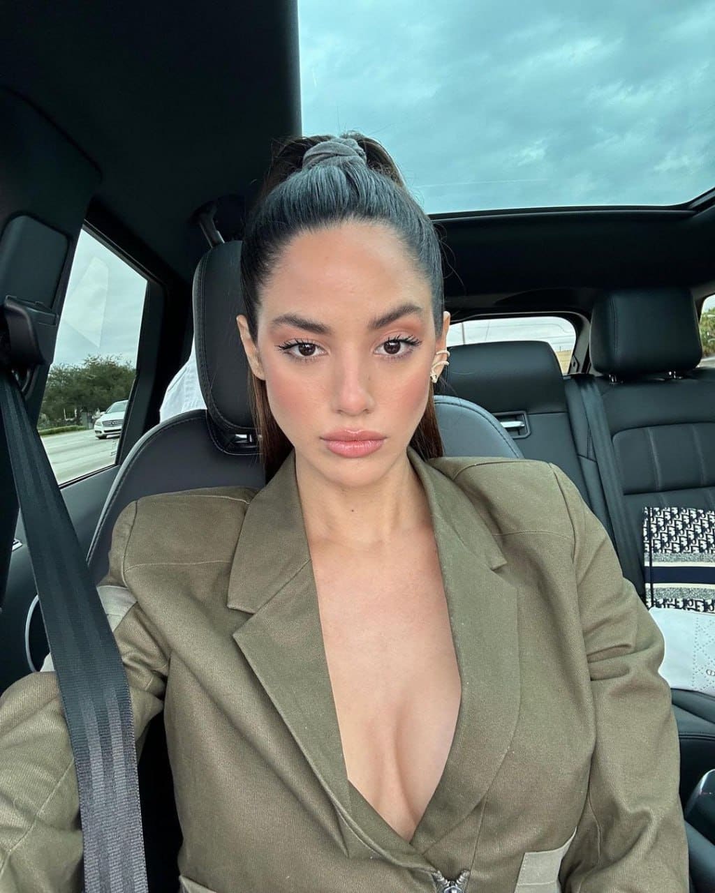 Emilia sits in a car back seat wearing an olive-green blazer with a deep V-neckline and a seatbelt across her chest.