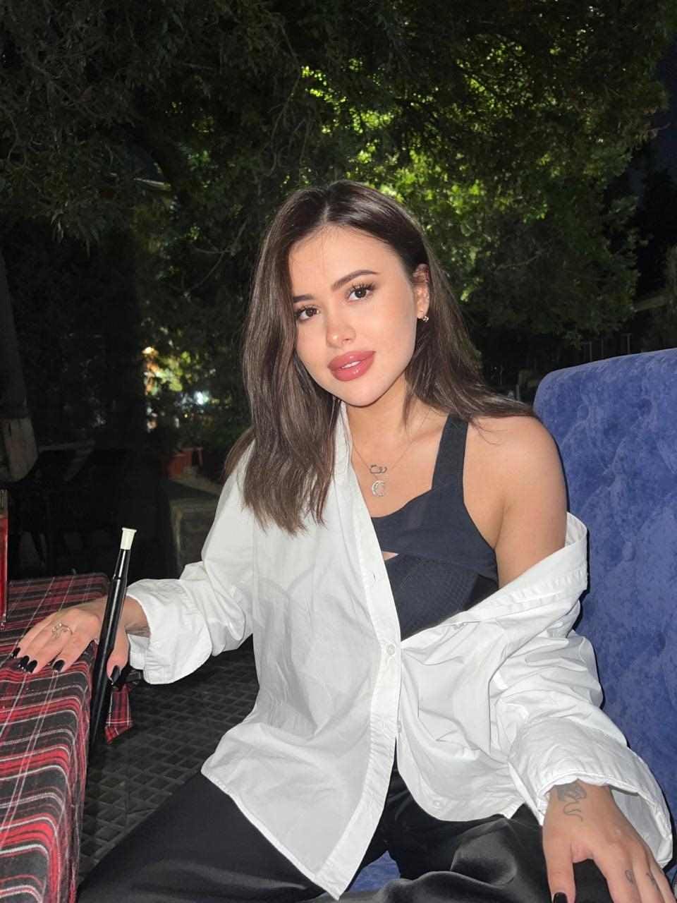 A woman in an open white shirt over a black tank sits at an outdoor table at night, on a blue chair, wearing a necklace.