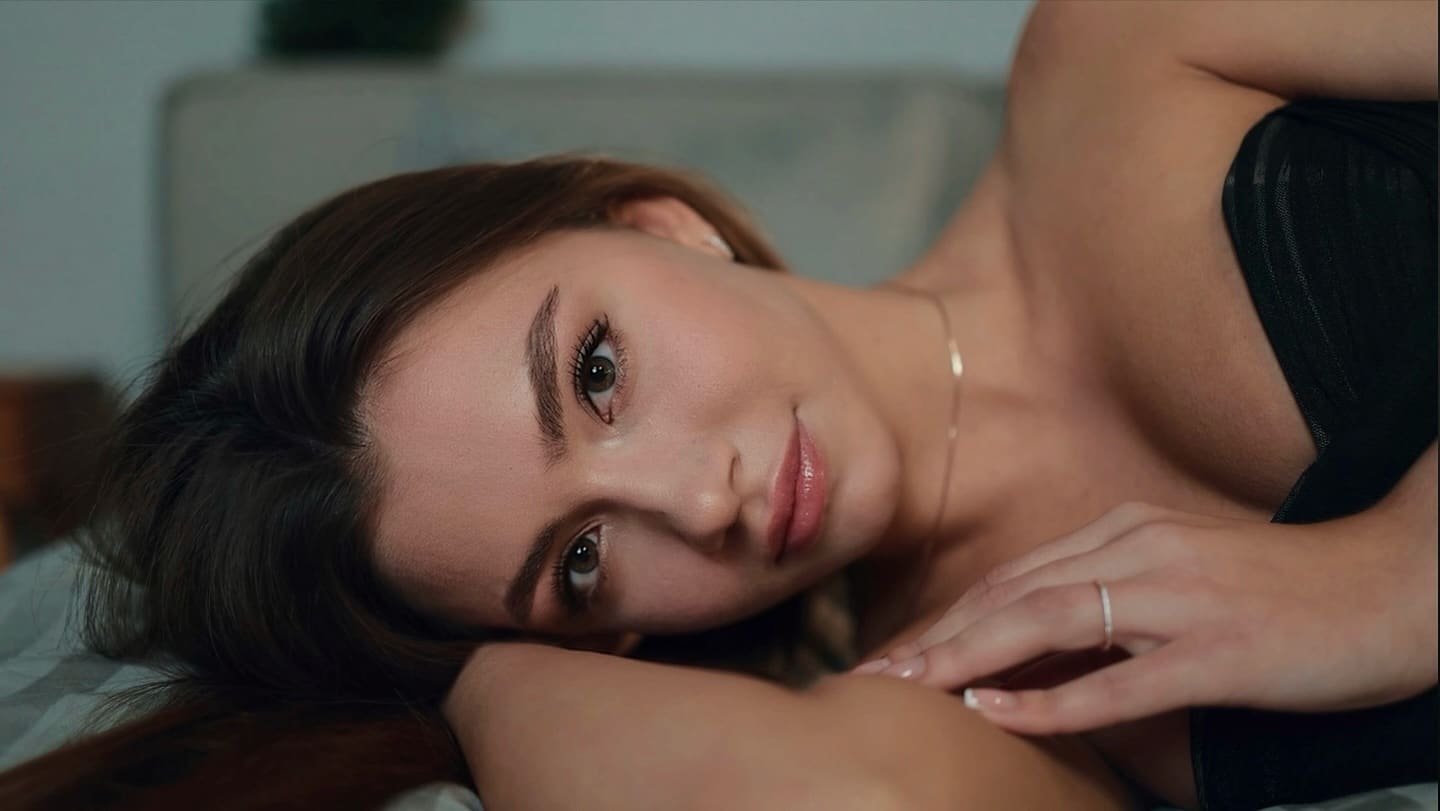 A woman in a black strapless top lies on her side on a bed, wearing a thin necklace with a hand near her shoulder.