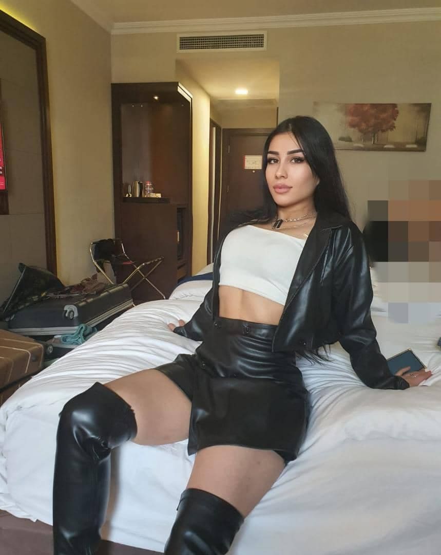 Marisa sits on a white hotel bed wearing a white crop top, black leather jacket, mini skirt, and thigh-high boots, with an open suitcase behind her.