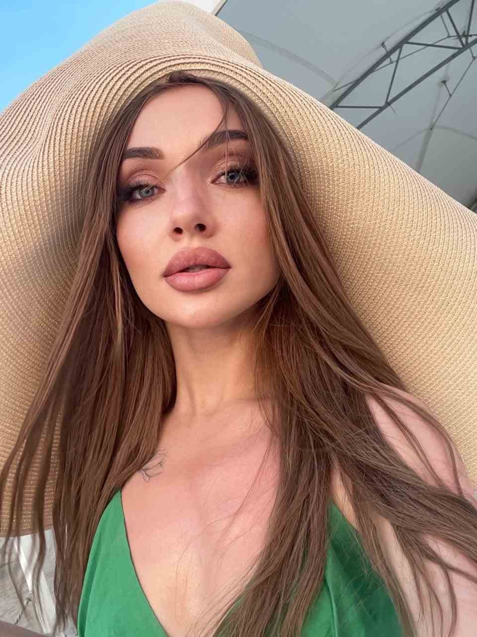 A woman wears a wide-brimmed straw hat and green V-neck top, posing outdoors under a bright sky with a metal structure in the background.