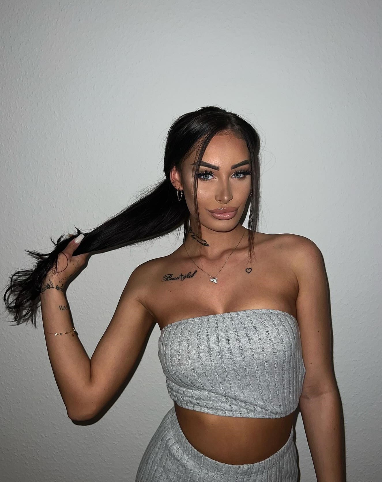 Maja stands against a white wall in a grey ribbed strapless crop top and high-waisted bottoms, holding her dark hair to one side.