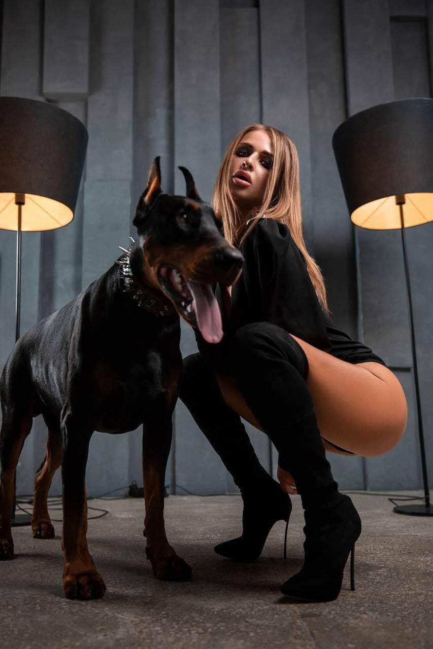 A blonde woman crouches beside a large dog in a room with grey paneled walls, wearing a black top, skirt, and thigh-high boots near two lamps.