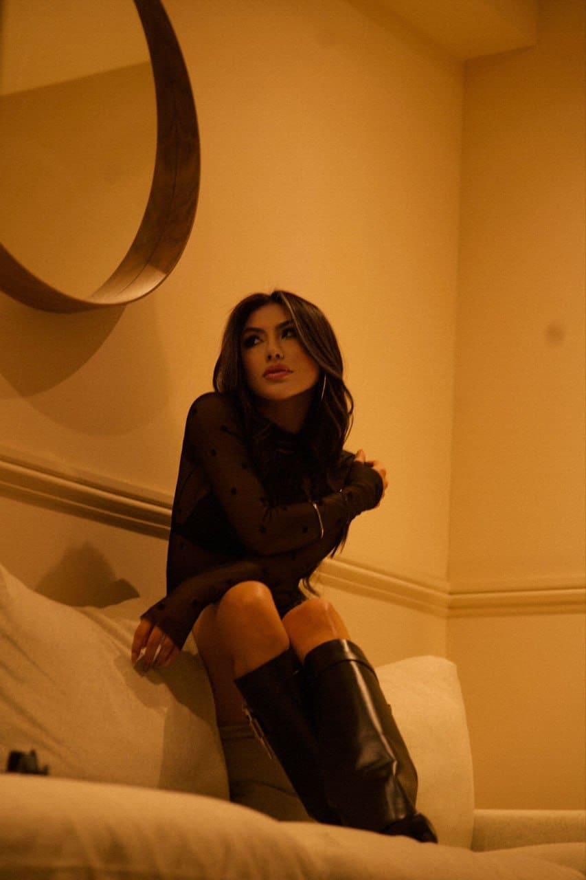 A woman sits on a sofa wearing a sheer black top and tall leather boots in a warmly lit room.