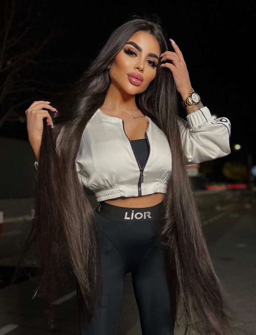 Mariam stands outdoors at night in a white cropped jacket and black leggings, holding her long dark hair.