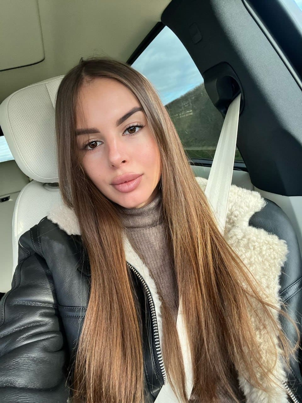 Sitting in a car passenger seat, she wears a black leather jacket with a shearling collar over a beige turtleneck, seatbelt fastened.