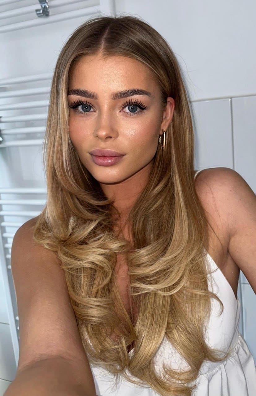 Emma with long wavy blonde hair wears a white sleeveless top and hoop earrings in a close-up selfie against white paneled walls.