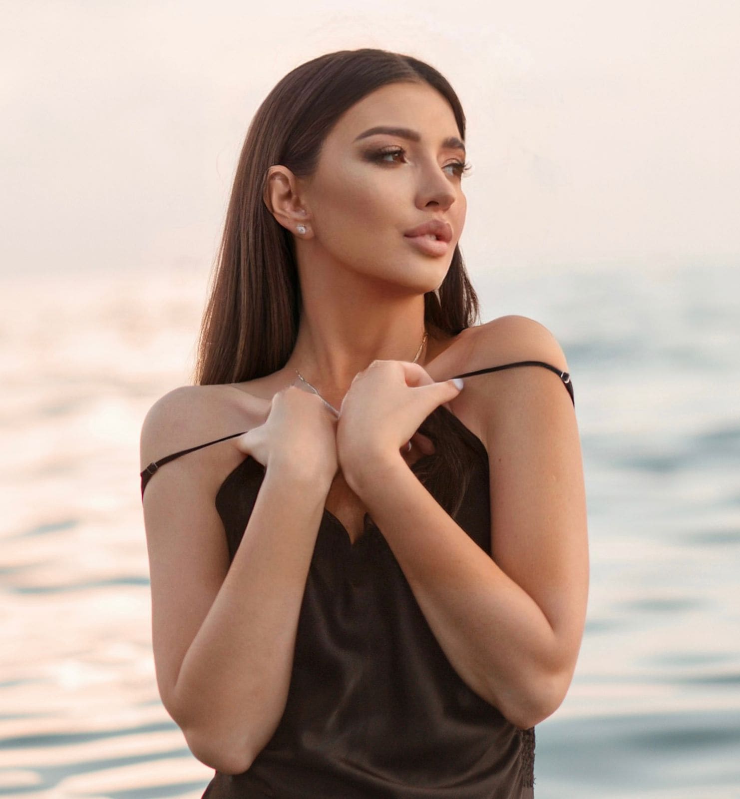 Aglaya stands by water in a black satin dress, pulling the straps off her shoulders to reveal bare skin.