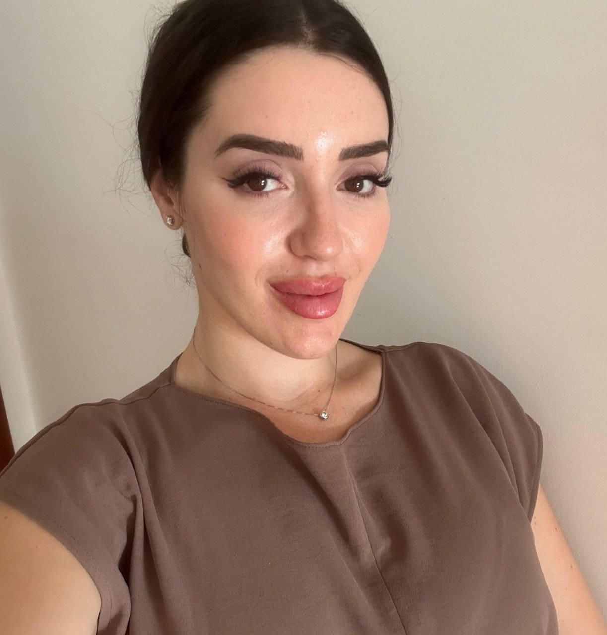 Marta takes a selfie against a plain light wall, wearing a brown short-sleeved top and thin necklace with dark hair pulled back.