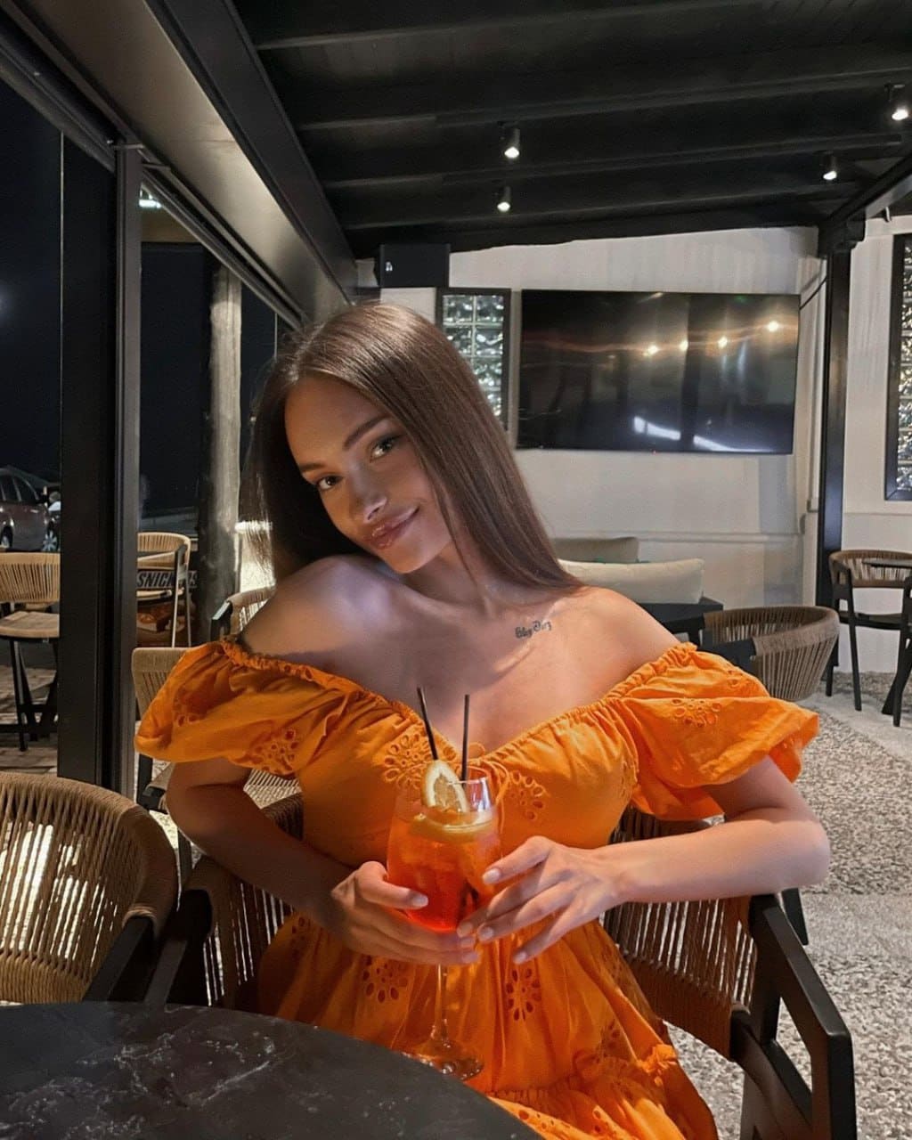 A woman in an orange off-the-shoulder dress sits at a dimly lit restaurant table, holding a tall glass with straws and citrus garnish.