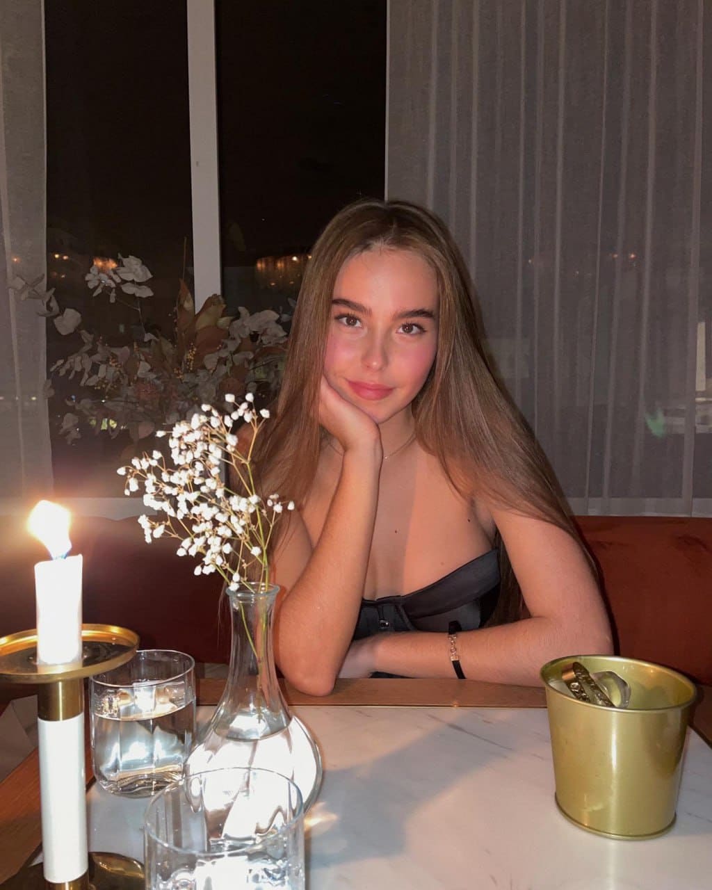 A woman in a dark strapless top rests her chin on her hand at a candlelit table with baby's breath flowers.